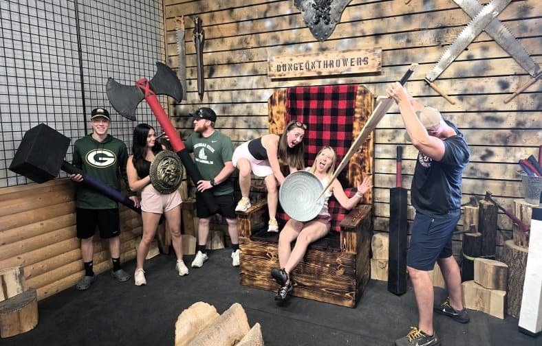 Six people posing with large foam weapons in front of a throne at Dungeon Throwers, with some playfully acting out a mock battle scene.