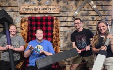 Group of four people posing with large foam weapons in front of a throne and a “Dungeon Throwers” sign at an axe throwing venue.